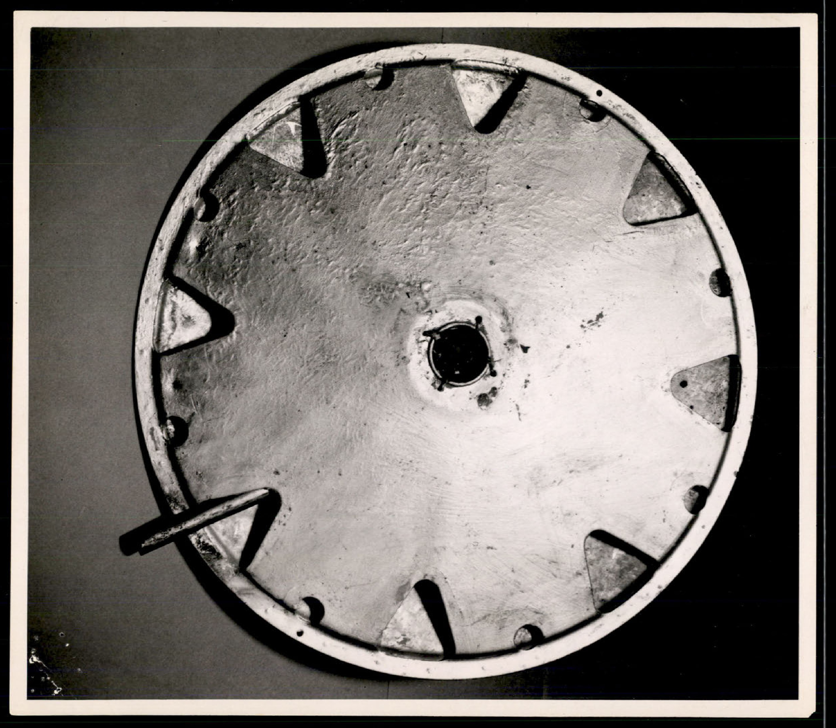Top or bottom view of a metallic, circular object with triangular cutouts around the perimeter and a central hole.