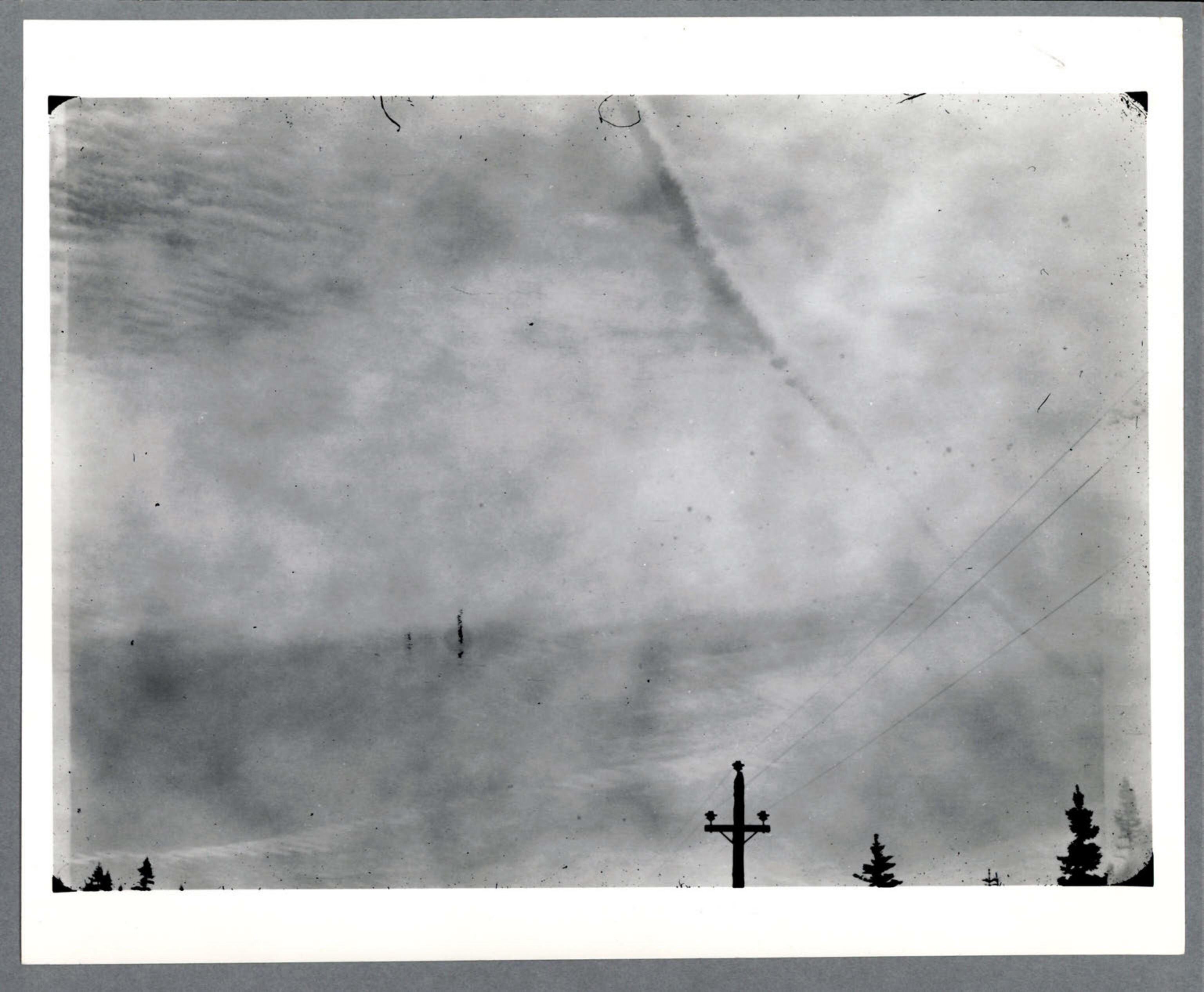 Photograph of a contrail in a cloudy sky, with power lines and trees in the foreground.