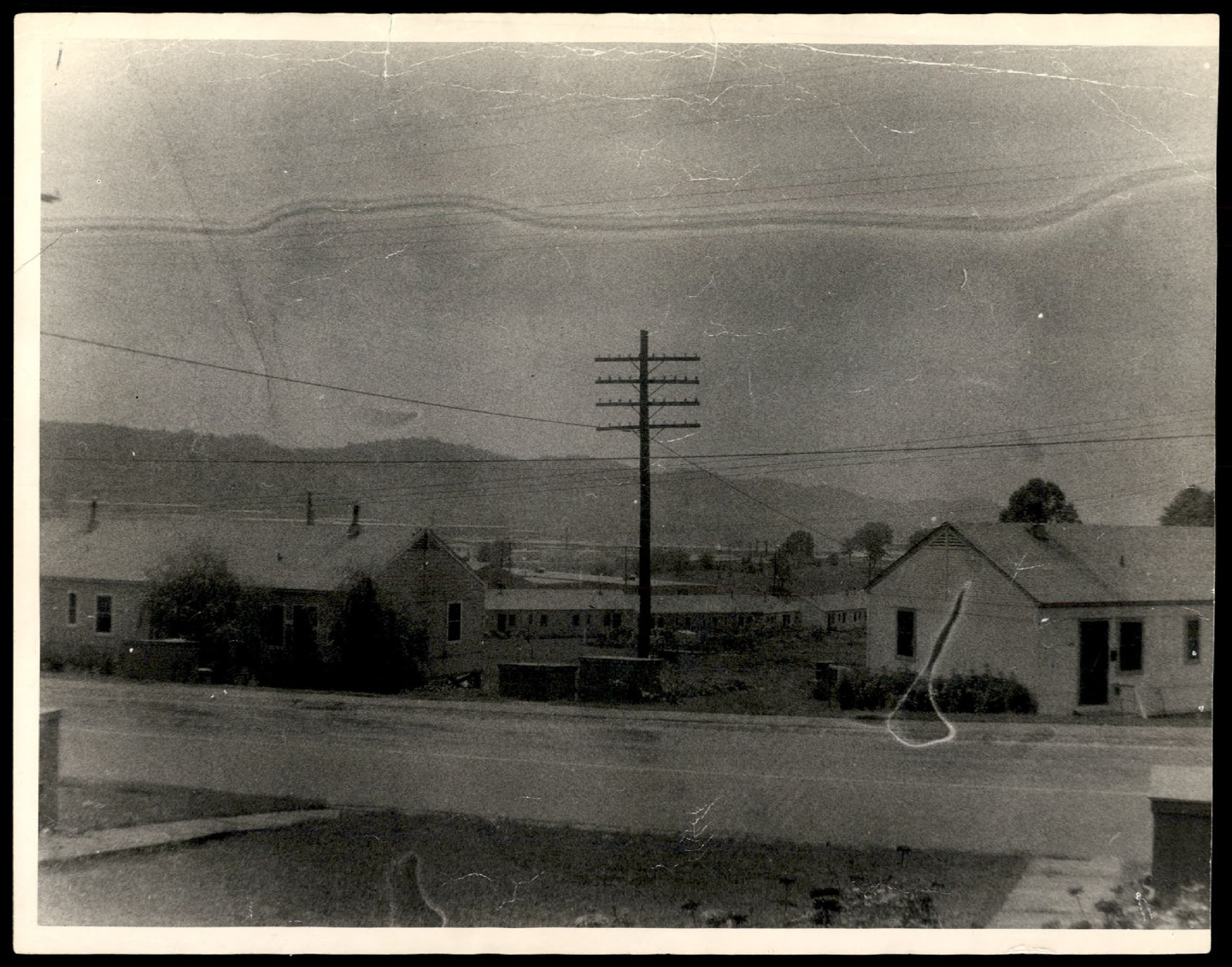 Photograph of a residential area with houses, a road, a telephone pole, and mountains in the background. This appears to be a photo of the scene without the anomalous object.